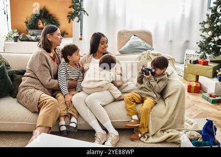 Ein asiatischer Junge, der ein Foto von der Familie macht, während er sich fröhlich in seinem modernen Wohnzimmer an einem wunderschön dekorierten Weihnachtsbaum sammelt. Stockfoto