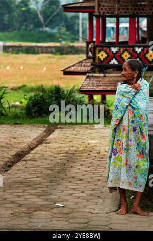 Eine alte Frau hüllt sich in eine Decke vor einem traditionellen Haus und Reisfeldern in Indonesien Stockfoto