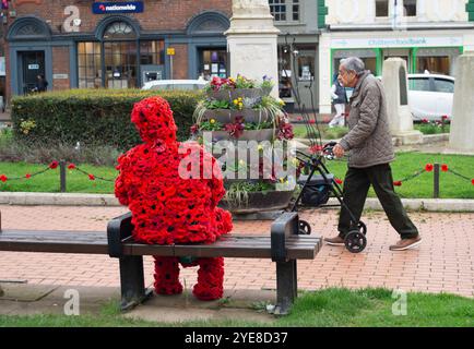 Chesham, Buckinghamshire, Großbritannien. Oktober 2024. Ein älterer Mann geht an einer auffälligen Figur des Mannes vorbei, der mit rotem Mohn bedeckt ist und auf einem Sitz in Chesham neben dem Kriegsdenkmal sitzt und eine weiße Taube hält. Quelle: Maureen McLean/Alamy Live News Stockfoto