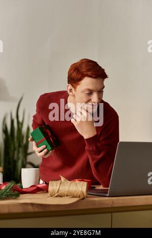 Ein fröhlicher junger Mann in gemütlicher Umgebung wickelt ein Weihnachtsgeschenk ein, während er seinen Laptop benutzt. Stockfoto