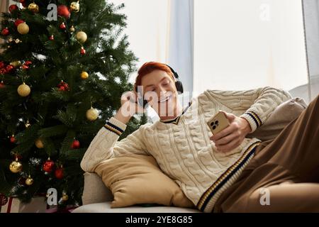 Ein fröhlicher junger Mann hört Musik über Kopfhörer, während er neben einem festlichen Weihnachtsbaum sitzt. Stockfoto