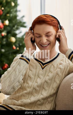 Ein entzückter junger Mann hört Musik, während er neben einem gut geschmückten Weihnachtsbaum sitzt. Stockfoto