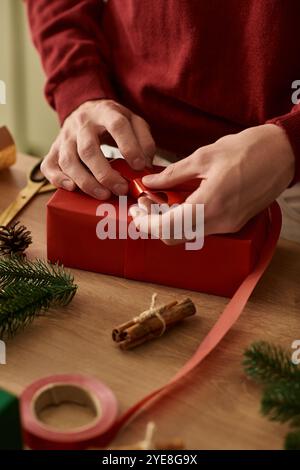 Ein junger Mann bindet vorsichtig ein Band um ein knallrotes Geschenk, während er sich auf Weihnachten vorbereitet. Stockfoto