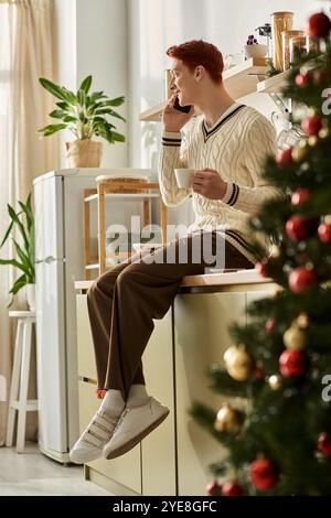 Ein junger Mann entspannt sich in seiner Küche, schlürft einen Drink und plaudert zu Weihnachten am Telefon. Stockfoto