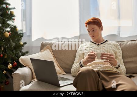 Der junge Mann genießt einen Drink auf einem gemütlichen Sofa neben einem wunderschön geschmückten Baum. Stockfoto