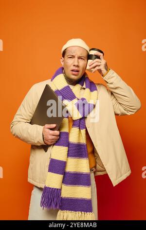 Ein junger Mann mit Tätowierungen balanciert einen Laptop und eine Kaffeetasse und trägt ein lebendiges Outfit. Stockfoto