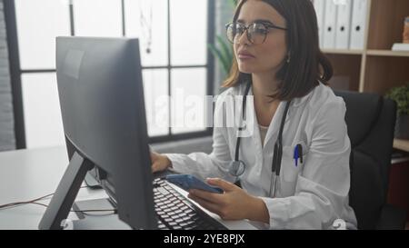 Hispanische Ärztin, die an einem Computer in einer Klinik arbeitet, ein Telefon hält, mit einem Stethoskop um den Hals. Stockfoto