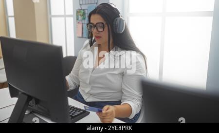 Frau mit Kopfhörern, die am Computer arbeitet und Kreditkarte in heller Büroumgebung hält Stockfoto