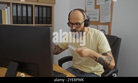 Hispanischer Mann in einem Büro, mit Kopfhörern und Brillen, am Computer, mit einem Dokument mit ernstem Ausdruck in einer organisierten Arbeitsplatte Stockfoto