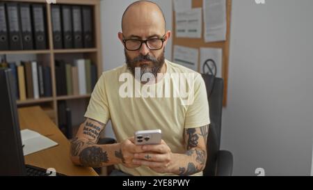 Ein hübscher hispanischer Mann mit Bart und Tattoos benutzt ein Smartphone in einem Büro mit Regalen und Dokumenten im Hintergrund Stockfoto