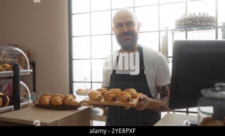 Hübscher hispanischer Mann mit Bart und Tattoos, der Gebäck in der Bäckerei mit Innenbeleuchtung hält Stockfoto