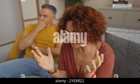 Ein Paar mittleren Alters hat eine ernsthafte Diskussion in ihrem Wohnzimmer, während die Frau Frustration ausdrückt, während der Mann nachdenkt Stockfoto