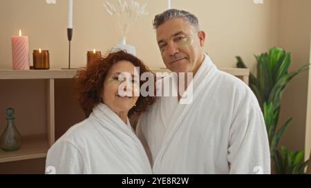 Paare mittleren Alters in weißen Bademänteln mit Augenmasken, die einen Spa-Tag drinnen mit beleuchteten Kerzen und Pflanzen für ein entspanntes Ambiente genießen Stockfoto