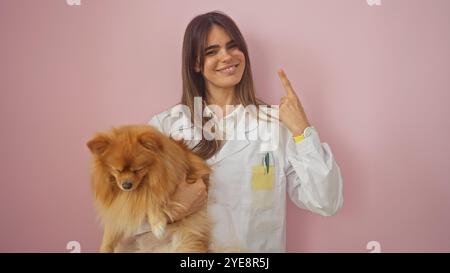 Eine junge erwachsene hispanische Frau in einem weißen Mantel, die einen pommerschen Hund vor einem rosa isolierten Hintergrund hält. Stockfoto