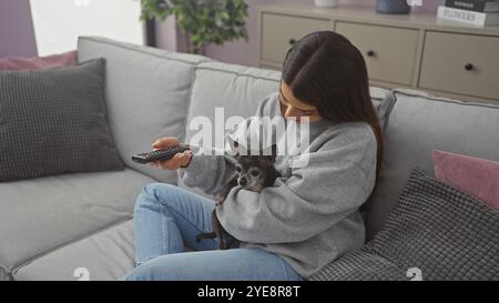 Eine junge Frau kuschelt sich mit ihrem chihuahua auf einem Sofa, während sie eine Fernbedienung in einem gemütlichen Wohnzimmer hält Stockfoto