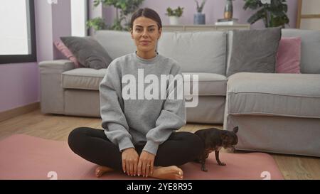 Eine junge Frau lächelt sanft, während sie im Kreuz mit ihrem Haustier chihuahua in einem gemütlichen Wohnzimmer sitzt. Stockfoto