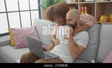 Interrassisches Paar kuschelt sich auf einem Sofa und schaut sich in einem gemütlichen Wohnzimmer einen Laptop an Stockfoto