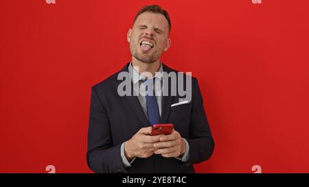Ein hübscher Mann im Anzug, der die Zunge ausstreckt, während er das Smartphone vor rotem Hintergrund hält. Stockfoto