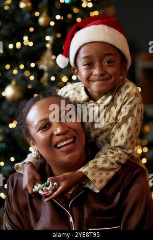 Vertikales Porträt der verspielten Mutter und des Sohnes, die gemeinsam Weihnachten feiern und mit einem Lächeln in die Kamera schauen, ein Junge mit Weihnachtsmütze Stockfoto