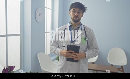 Ein junger arabischer Mann in einem weißen Mantel mit Stethoskop und Tablette steht in einem Krankenhauszimmer. Stockfoto