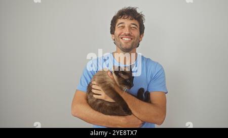 Ein glücklicher hispanischer Mann, der eine siamesische Katze an einer weißen Wand umarmt Stockfoto