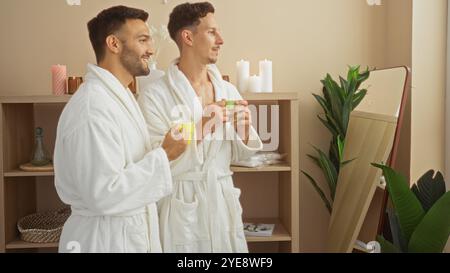 Zwei Männer in weißen Bademänteln genießen Getränke in einem mit Kerzen und Pflanzen geschmückten Spa-Raum, der ein liebevolles Paar in einem friedlichen Wellness-Bereich repräsentiert Stockfoto