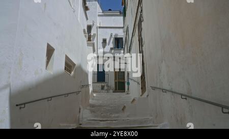 Schmale, weiß getünchte Gasse mit Stufen in ostuni, apulien, italien, die zu traditionellen Häusern unter einem klaren blauen Himmel mit mediterranem Bogen führt Stockfoto