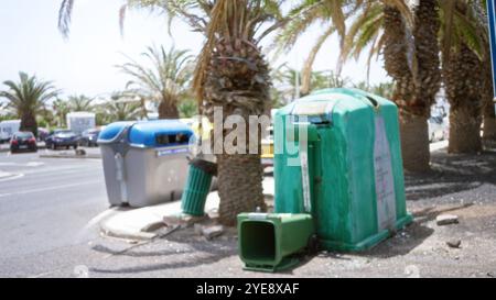 Defokussierte Abfallbehälter in einem sonnigen Stadtgebiet mit verschwommenen Palmen im Hintergrund, die Recyclingbehälter und Müll auf dem Boden hervorheben. Stockfoto