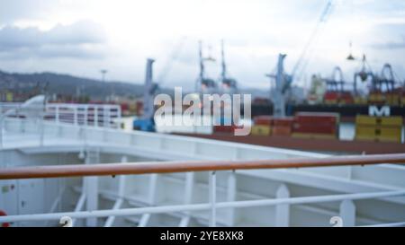 Unscharfe Sicht auf einen Frachthafen mit Containern und Kränen unter bewölktem Himmel, während Sie auf dem Deck eines Schiffes stehen. Stockfoto