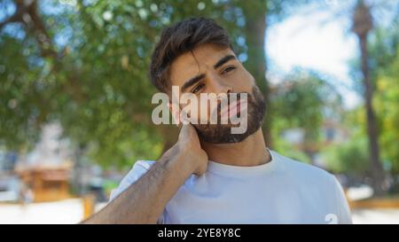 Junger Mann mit Bart, der seinen Hals vor Schmerzen berührt, in einem urbanen Park im Freien Stockfoto