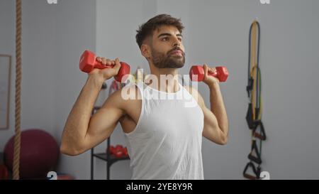 Gutaussehender Mann, der drinnen mit Kurzhanteln in einer Turnhalle tragt, ein weißes ärmelloses Hemd trägt und sich mit einem entschlossenen Ausdruck auf sein Workout konzentriert Stockfoto