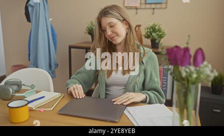 Eine junge blonde Frau in Brille, die an einem Laptop arbeitet, in einem gemütlichen Heimbüro mit Pflanzen und Büchern. Stockfoto