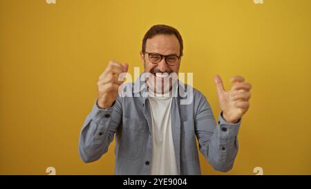 Hispanischer Mann mittleren Alters, der fröhlich mit geballten Fäusten vor einem leuchtend gelben Hintergrund lächelt und ein reifes, schönes Aussehen mit g zeigt Stockfoto