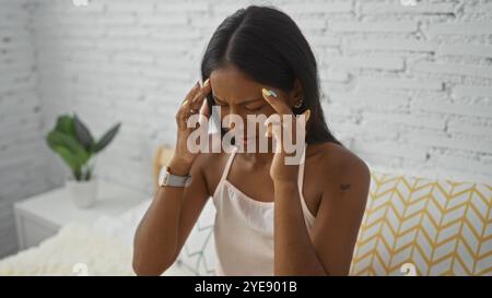 Frau, die unter Kopfschmerzen leidet, während sie auf dem Bett sitzt, in einem modernen Schlafzimmer mit weißem Backsteinmauerhintergrund Stockfoto