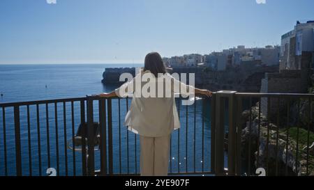 Eine junge hispanische Frau bewundert den wunderschönen Meerblick von einem Aussichtspunkt in polignano a Mare, apulien, italien, unter einem klaren blauen Himmel und umgeben von Stockfoto