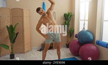 Junger, gutaussehender, hispanischer Mann, der sich in einem Fitnessraum mit Fitnessgeräten und Pflanzen in Innenräumen ausdehnt und in einem modernen s eine Fitnessroutine präsentiert Stockfoto