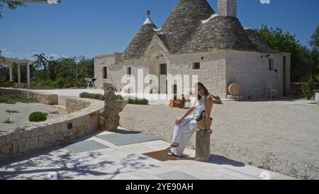 Junge hispanische Frau, die in der Nähe des traditionellen Trullihauses in alberobello, apulien, italien sitzt und den Sommertag in der Altstadt, umgeben von Historie, genießt Stockfoto