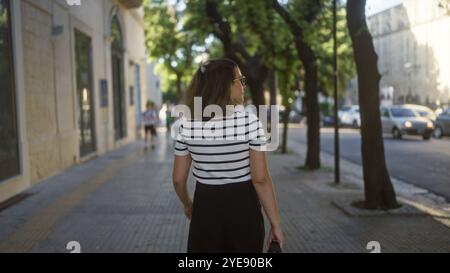 Eine junge, schöne hispanische Frau läuft an einem sonnigen Tag eine von Bäumen gesäumte Straße in der charmanten Stadt lecce, apulien, italien. Stockfoto