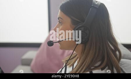 Eine junge Frau mit brünetten Haaren, die ein Headset trägt, arbeitet in Innenräumen ferngesteuert und veranschaulicht die Umgebung des Home Office. Stockfoto