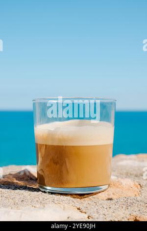 Ein Café Latte oder ein Latte Macchiato in einem Glas, auf einer rustikalen Stein- und Mörsermauer, vor dem Meer, mit etwas freiem Platz am Himmel Stockfoto