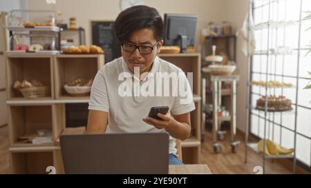 Hübscher junger Mann mit Brille und weißem Poloshirt mit Smartphone und Laptop in einer gemütlichen Bäckerei mit verschiedenen Backwaren in den Ausstellungsregalen Stockfoto