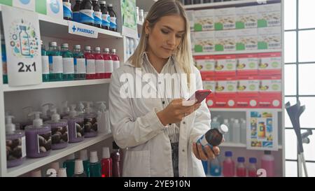 Eine junge, blonde Frau in einer Apotheke überprüft ihr Handy, während sie eine Flasche hält, umgeben von Regalen mit bunten Produkten. Stockfoto