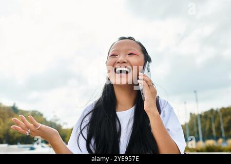 Eine stilvolle junge asiatische Frau mit langen Haaren lacht fröhlich, während sie am Telefon spricht und das Leben im Freien annimmt. Stockfoto