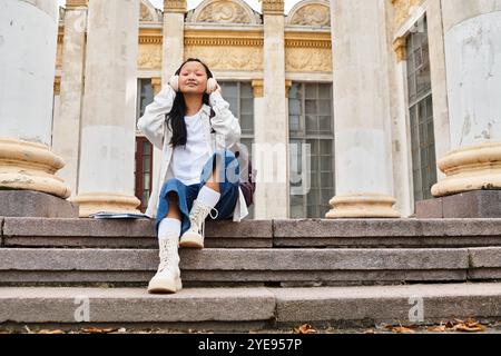 Eine stilvolle junge Studentin entspannt sich auf den Stufen der Universität, verloren in ihrer Musik und genießt das pulsierende Leben auf dem Campus. Stockfoto