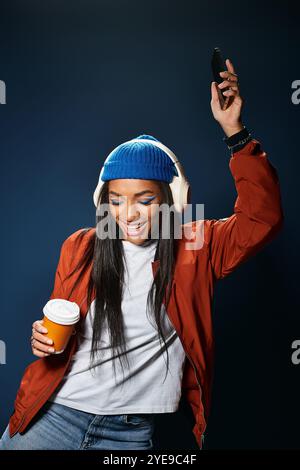 Eine stilvolle junge Frau mit Kopfhörern tanzt fröhlich, während sie Kaffee in einem gemütlichen Herbstlook hält. Stockfoto