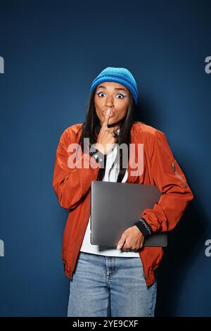 In einem stilvollen, warmen Outfit gibt eine junge Frau spielerisch Gesten, während sie einen Laptop hält. Stockfoto