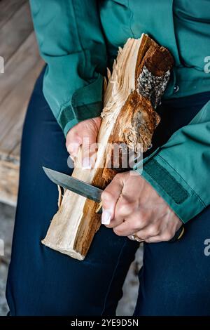 Nahaufnahme weiblicher Hände, die geschickt einen Federstock aus Spaltholz schnitzen. Eine Frau, die ein Überlebensmesser benutzt und sich auf ein Lagerfeuer im Freien vorbereitet. Stockfoto