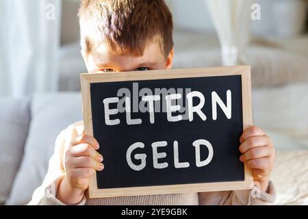 Bayern, Deutschland - 30. Oktober 2024: Kind, das eine Tablette in der Luft hält, mit Text ELTERNGELD. Symbolisches Bild für Zahlungen und Antrag der Eltern auf staatliches Geld und finanzielle Freistellung der Familien durch Zahlung. PHOTOMONTAGE *** Kind hält eine Tafel in die Luft, mit Text ELTERNGELD. Symbolbild für Zahlungen und Antrag für Eltern auf Geld vom Staat und finanzielle Entlassung der Familien durch Auszahlung. FOTOMONTAGE Stockfoto