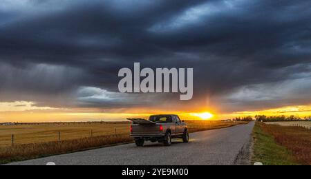 Der Pickup Truck mit großer Ladung fährt bei Sonnenuntergang durch die Landschaft Albertas mit dunklen Gewitterwolken über der Straße; Alberta, Kanada Stockfoto