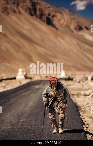 Senior man läuft mit einem Stock auf einer Asphaltstraße in der Nähe von Kurgiakh im Zanskar-Tal, Ladakh, Indien; Zanskar-Tal, Ladakh, Indien Stockfoto